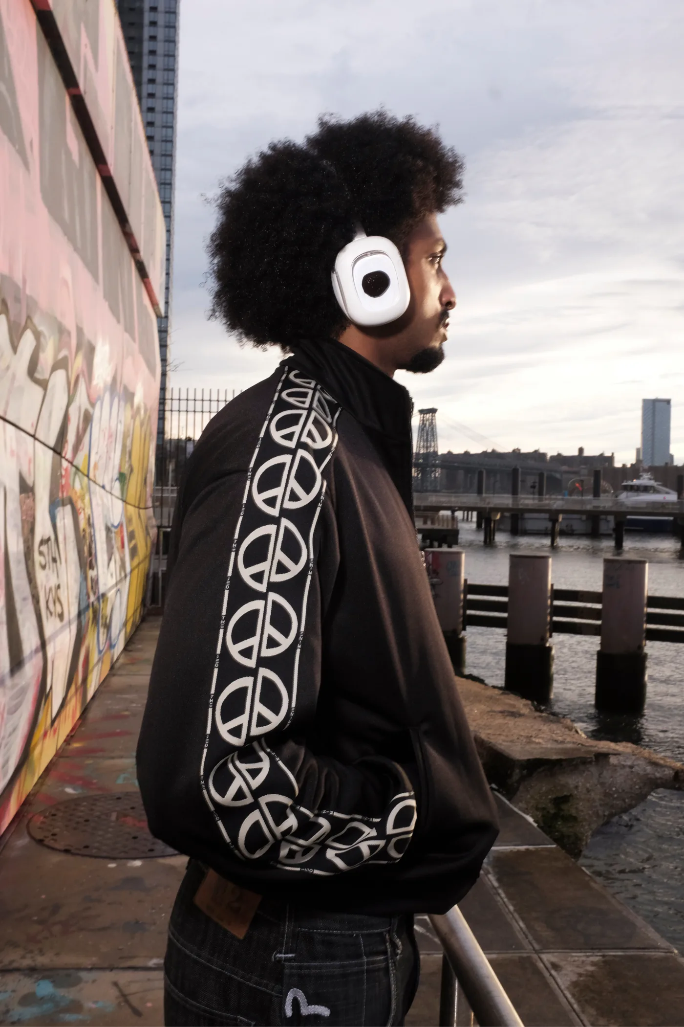 Man in a black peace-sign track jacket wearing white Volta ferrofluid headphones by the East River